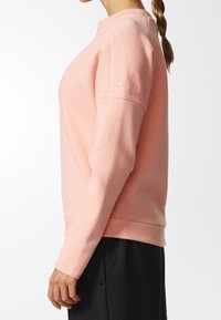 Mujer vestida con una sudadera de manga larga Adidas rosa claro y pantalones negros texturizados, mostrada de lado contra un fondo blanco liso.