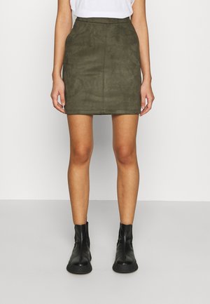 Kvinde iført en mørkegrøn ruskind mini nederdel med en hvid top og sorte ankelstøvler, stående mod en ensartet lys baggrund.