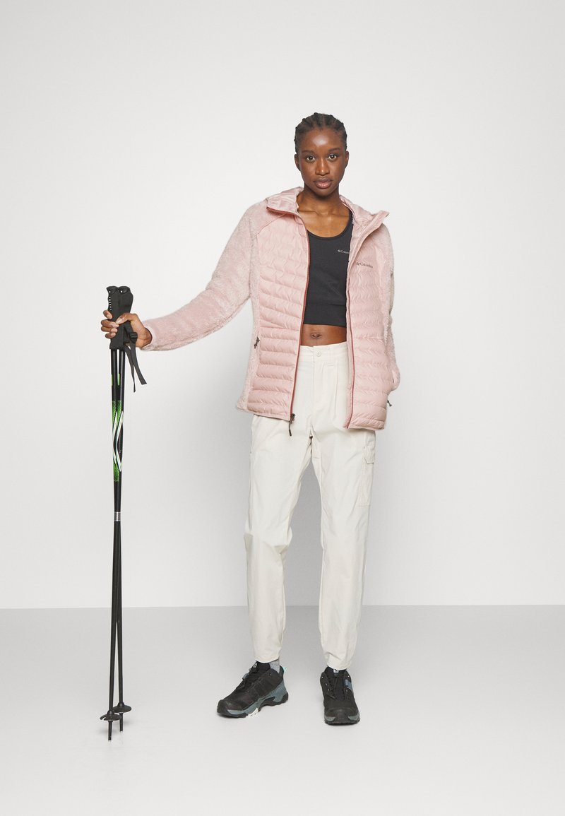 Frau in hellrosa Jacke, schwarzem Crop-Top, weißen Hosen und schwarzen Wanderschuhen, die schwarze und grüne Trekkingstöcke hält.