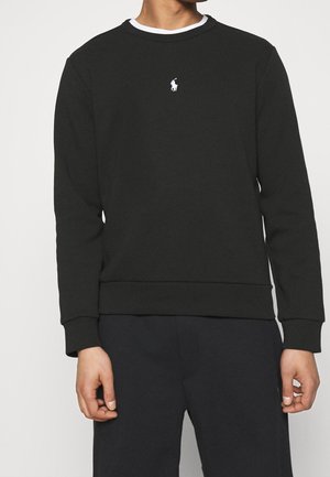 Man draagt een zwarte trui met lange mouwen met een wit polo-spelerlogo op de borst, gecombineerd met zwarte broek, staand met ontspannen armen.