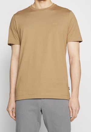 Mann trägt ein einfarbiges kurzärmeliges hellbraunes T-Shirt mit kleinem "BOSS"-Logo auf der Brust und graue Hose vor weißem Hintergrund.