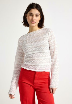 Ung kvinde med kort mørkt hår iført en hvid tekstureret bluse med lange ærmer og højtaljede, knaldrøde bukser, stående mod en ensfarvet baggrund.