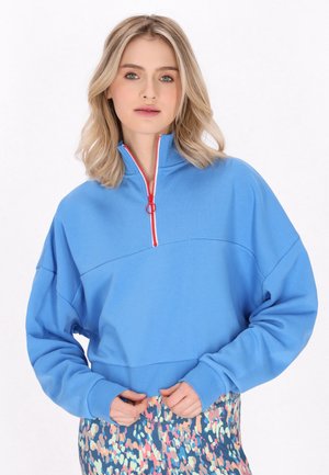 Ung kvinde med blondt hår iført en blå trøje med kvart lynlås og flerfarvede, mønstrede leggings, stående mod en ensfarvet hvid baggrund.