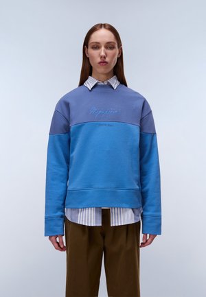 Junge Frau trägt ein mehrlagiges blau gestreiftes Hemd und einen zweifarbigen blauen Napapijri-Sweatshirt mit brauner Hose, steht vor einem schlichten hellen Hintergrund.