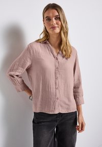 Frau mit langen blonden Haaren, die Creolen, eine hüftlange knöpfbare Bluse in Mauve und schwarze Jeans trägt, steht vor einer weißen Wand.
