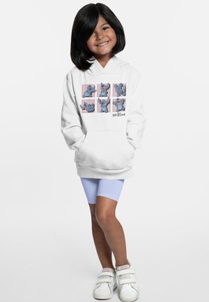 Weiße Kapuzenjacke mit einem grafischen Design, das blaue Cartoon-Figuren auf einem rosa Hintergrund zeigt. Das Model trägt hellblaue Shorts und weiße Schuhe.