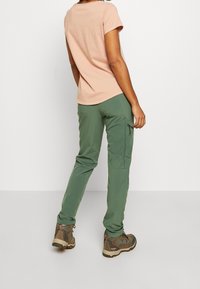 Lichtroze T-shirt met korte mouwen, gecombineerd met groene stretch cargo broek met ritszakken, en bruine wandelschoenen met rode accenten.