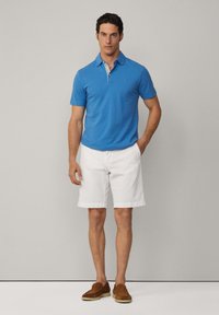 Blå pikétröja med krage, vita shorts och bruna slip-on skor. Modellen står med händerna i fickorna, neutral bakgrund.