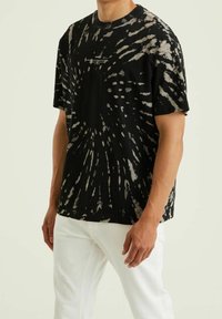 Muž v černo-pískovém tričku s krátkým rukávem s tie-dye vzorem a bílých kalhotách, stojící na pozadí s jednoduchou světle barevnou stěnou.