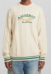 Krämfärgad tröja med kabelstickat mönster, rund krage samt ribbade ärmslut och nederkant i gröna ränder. Broderad text "AMHERST", 1998.