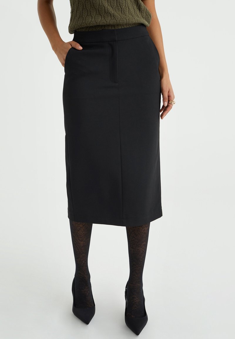 Jupe crayon noire avec une texture lisse, dotée d'une poche avant et d'une coupe au niveau des genoux, associée à des collants en dentelle noirs et des escarpins pointus.