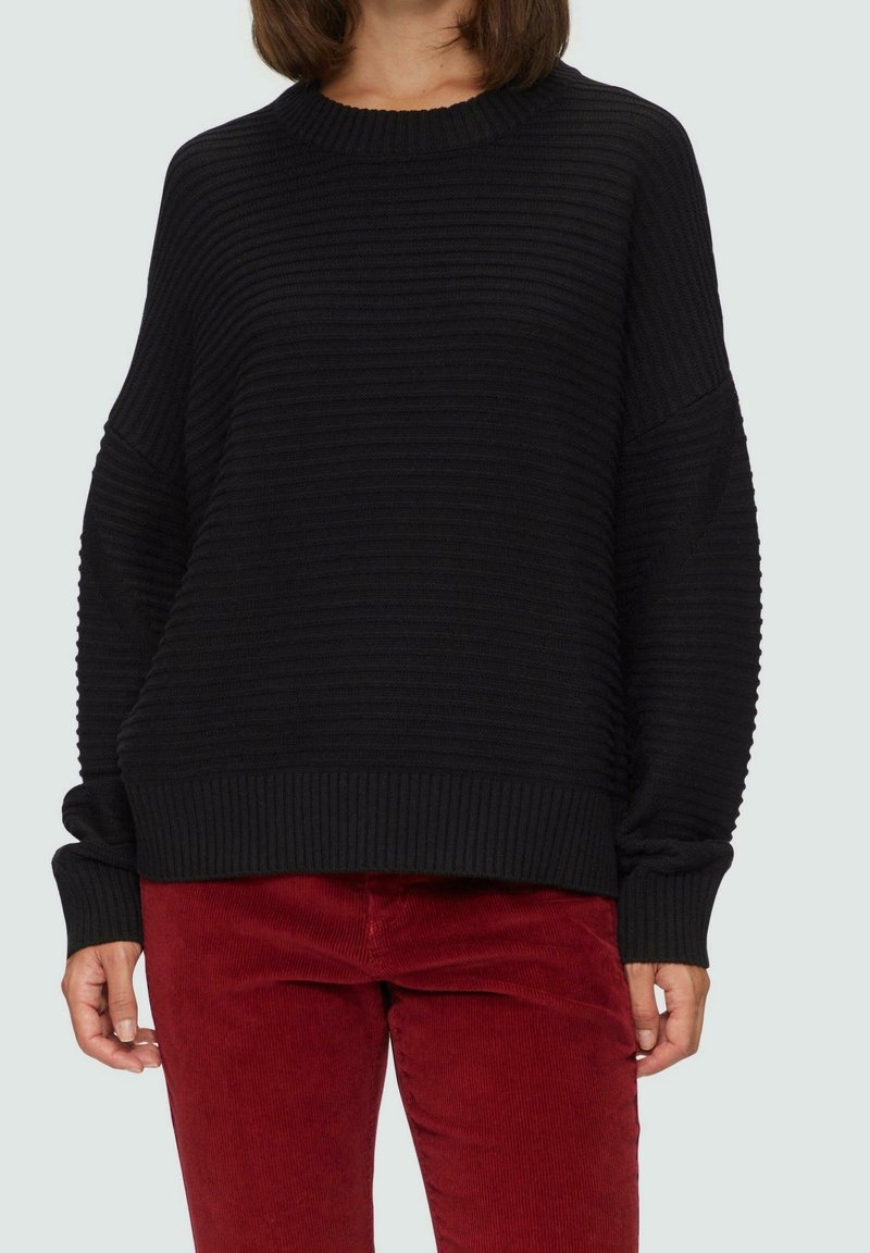 Personne portant un pull noir côtelé ample et un pantalon en velours côtelé rouge, debout devant un fond clair uni.