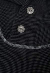 Schwarzer strukturierter Stoff mit zwei grauen Knöpfen und weißem gesticktem Rand auf einem gefalteten Kleidungsstück.