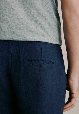 Személy sötétkék nadrágot visel hátul paszpólos zsebbel és egy szürke melírozott inget, látható a derék és a jobb kar oldala.