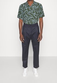 Camicia corta a maniche corte floreale verde con motivi bianchi, abbinata a pantaloni blu scuro e sneakers bianche. Taglio pulito e vestibilità sartoriale.
