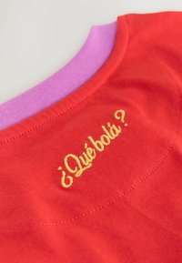 Slojevi crvene i ljubičaste tkanine sa žutim izvezenim tekstom „¿Qué bola?“ na crvenoj tkanini blizu šava.