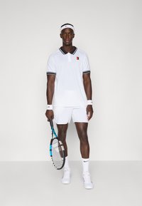 Jugador de tenis masculino con un uniforme blanco sosteniendo una raqueta de tenis negra y azul, de pie contra un fondo claro y uniforme.
