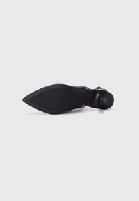 Μαύρο γυναικείο παπούτσι με μυτερή μύτη και ψηλό τακούνι, με υφασμένο ελαστικό πέλμα και λουράκι στον αστράγαλο με χρυσή αγκράφα, απεικονίζεται από κάτω σε λευκό φόντο.