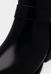 Svart lackläderankelboots med en slät, glansig yta, en bred rem runt vristen och en fyrkantig tåform.