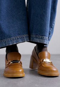 Mocasines de tacón en piel color cuero con un tacón grueso, que presentan un accesorio de cadena dorada y una suela texturizada, combinados con pantalones de mezclilla anchos.