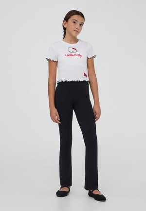 Top bianco a maniche corte con bordi arricciati neri, caratterizzato da una grafica rossa di "Hello Kitty"; abbinato a leggings neri aderenti e scarpe nere.