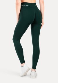 Leggings verde a vita alta realizzati in tessuto elastico, con una texture liscia e un dettaglio logo sulla cintura, abbinati a sneakers bianche.