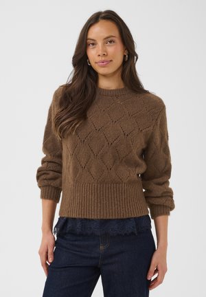 Femme aux longs cheveux bruns portant un pull en maille à motifs bruns, un jean bleu foncé et des créoles dorées, debout devant un fond uni.