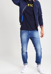 Marinblå zip-up jacka med randig krage, blå detaljer; svart t-shirt med gul text; ljusblåjeans; vita sneakers.