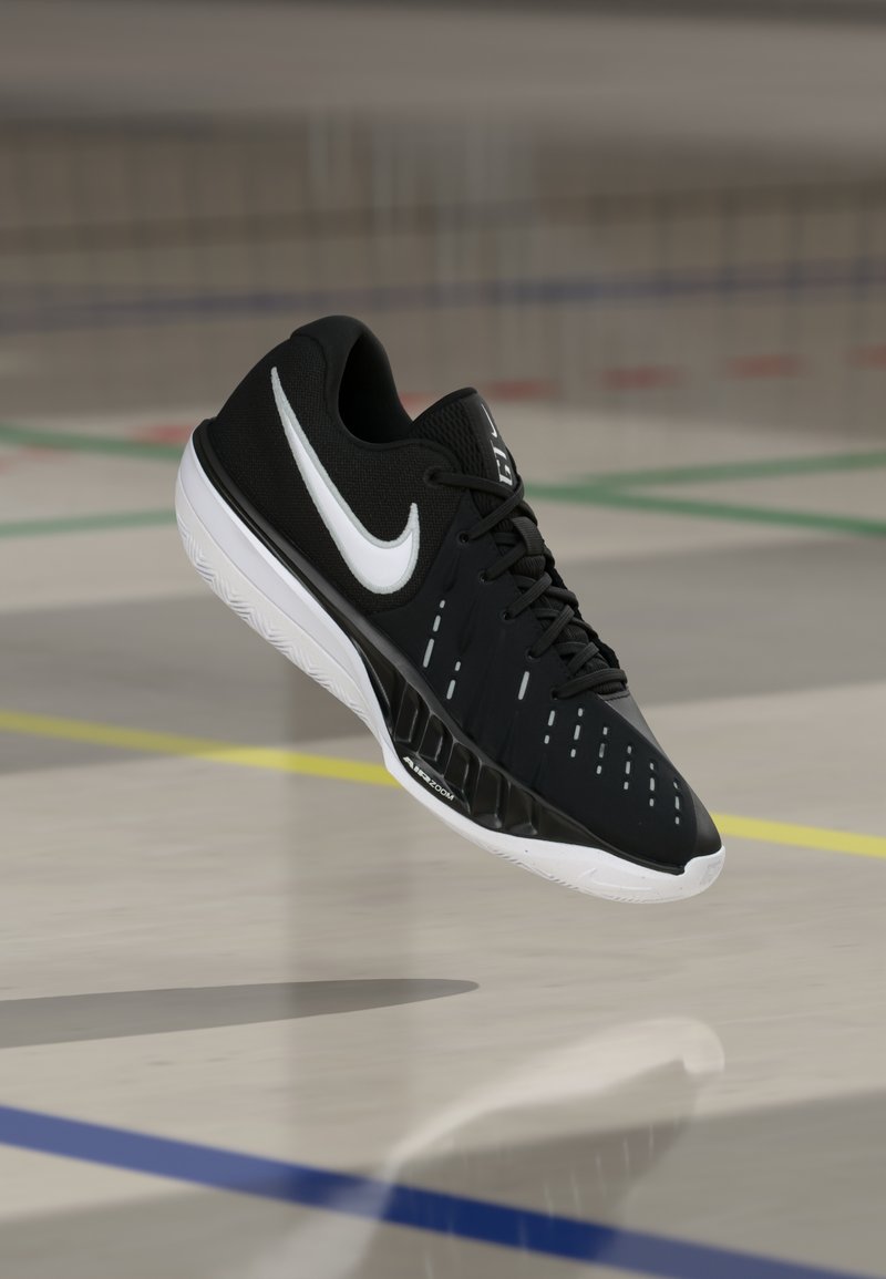 Zapatilla deportiva Nike Air Zoom en blanco y negro suspendida sobre un suelo de gimnasio marcado con líneas de colores.