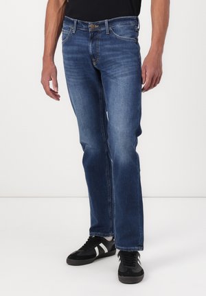 Mann iført rett blå jeans og svarte joggesko med hvite striper, stående mot en ensfarget hvit bakgrunn.