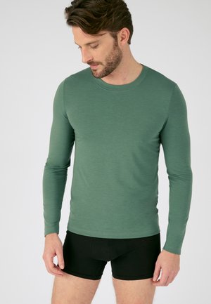 Camisa de manga larga en verde apagado, hecha de una tela suave, con cuello redondo. Combinada con pantalones cortos ajustados negros. Diseño minimalista sin patrones.