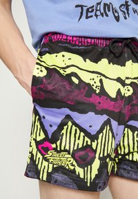 Person trägt bunte Netzshorts mit abstrakten Mustern und "Street Fighter"-Logo, kombiniert mit einem teilweise sichtbaren blauen Shirt.