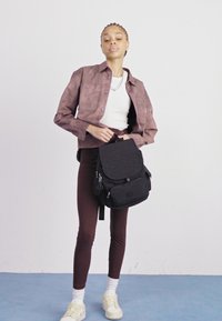Black backpack with zippered compartments and a flap, worn on one shoulder. Model in brown leggings, a white top, and a brown jacket.