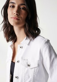 Chaqueta de mezclilla blanca con cierre de botón, herrajes plateados y bolsillos en el pecho. Diseño estructurado con costuras mínimas y un acabado limpio.