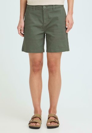 Mujer vistiendo pantalones cortos hasta la rodilla de color verde oliva y sandalias marrones con doble hebilla, de pie frente a un fondo liso.