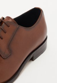 Scarpa di pelle marrone con una superficie liscia, punta rotonda e design tradizionale con lacci. Presenta un tacco basso impilato e cuciture minime.