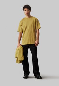 T-shirt oversize giallo senape in cotone, vestibilità rilassata, maniche corte. Il modello tiene un maglione giallo peloso. Pantaloni neri e scarpe completano il look.