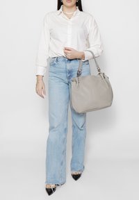 Sac à main en cuir gris avec une texture douce et un matériel argenté, présenté aux côtés d'un mannequin portant une chemise blanche et un jean bleu.