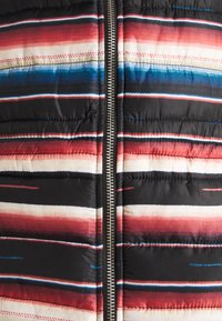 Vuorattu liivi, jossa on musta pohja ja vaakaraitoja punaisena, sinisenä ja kermaisen valkoisena. Keskellä kulkeva hopeinen vetoketju, ja pinnassa on quiltattu rakenne.