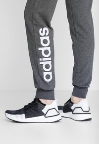 Zwart-witte sportschoenen met een gestructureerd bovenwerk, gecombineerde materialen en een stevige zool. Grijze joggingsbroek met groot wit "adidas" logo.