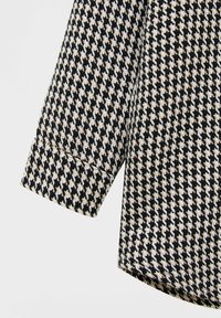 Zwart-wit stof met een hondenbeetpatroon en een gladde textuur. Voorzien van een gerolde mouw en een rechte zoom.