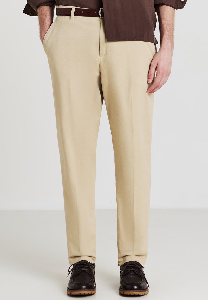 Pantaloni beige dritti indossati con cintura e scarpe in pelle marrone scuro, abbinati a una camicia marrone scuro, con una mano in tasca.