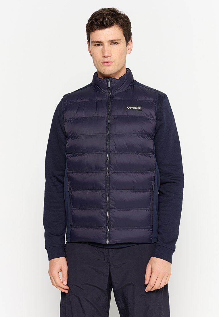 Gilet rembourré bleu marine avec un col montant, fermeture éclair sur le devant et détail de logo. Texture lisse avec des sections matelassées et poches latérales.