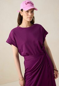 Robe violette à manches courtes avec un détail de taille froncée. Associée à une casquette rose portant le mot "AMORA" en lettres majuscules. Texture du tissu lisse.