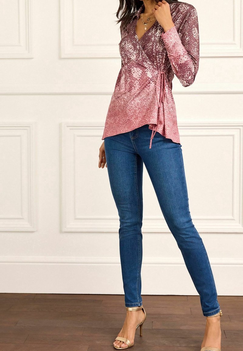 Blusa a portafoglio rosa con pattern texturizzato, abbinata a jeans aderenti blu e sandali dorati con fasce. Tessuto morbido e vita definita.