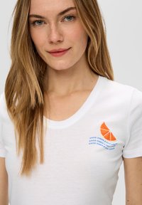 Witte katoenen t-shirt met een ronde halslijn en korte mouwen. Geborduurd graphic van een sinaasappel met blauwe tekst "GOEDE TIJDEN & TAN LINES."