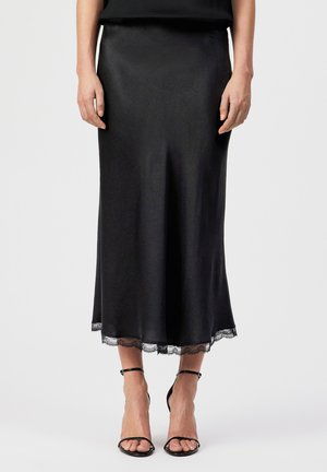 Donna che indossa una gonna midi nera con bordo in pizzo all'orlo e sandali neri con cinturino alla caviglia e tacco alto, in piedi davanti a uno sfondo semplice.