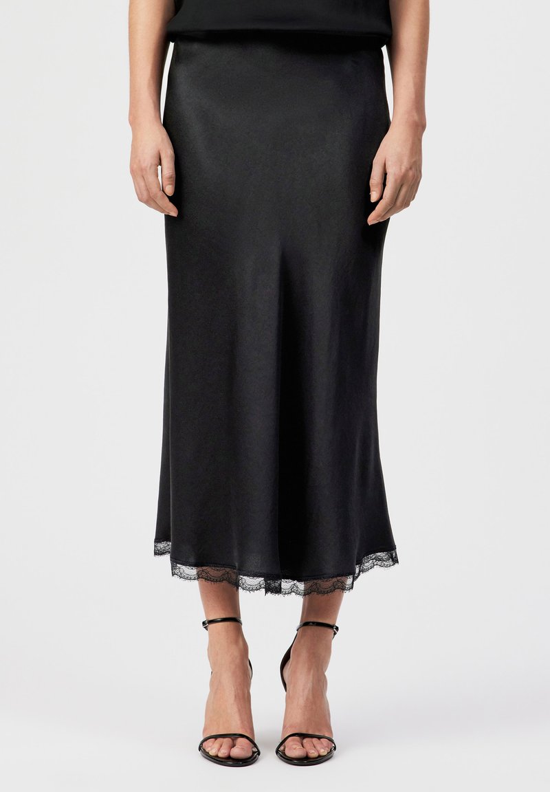 Femme portant une jupe midi noire bordée de dentelle à l'ourlet et des sandales à talons hauts avec bride à la cheville noires, se tenant devant un fond uni.