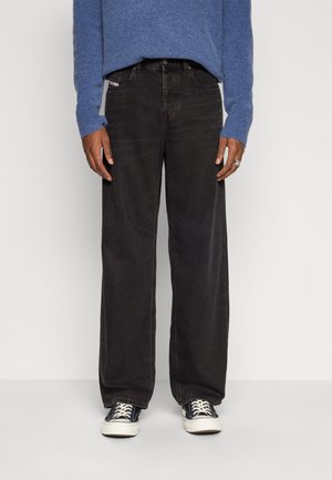 Person iført sorte jeans med vide ben, blå langærmet sweater, sorte og hvide sneakers, stående mod en ensfarvet hvid baggrund.