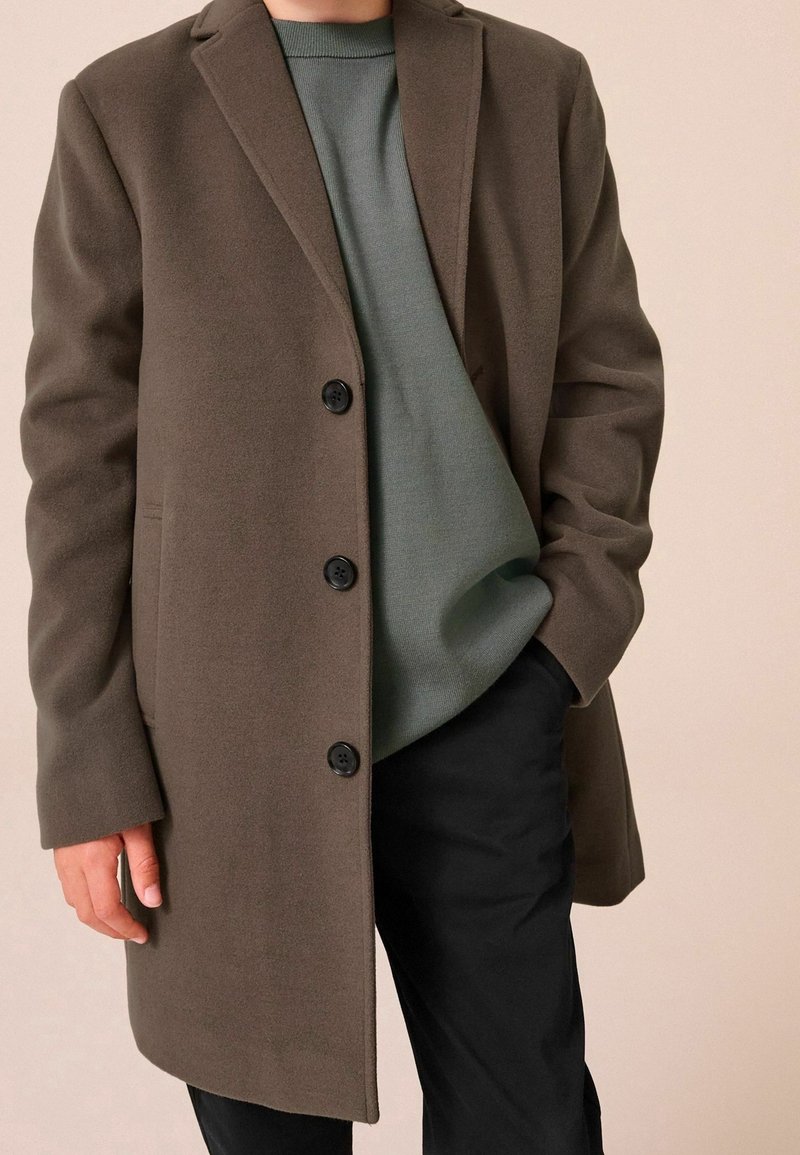 Personne portant un manteau marron boutonné par-dessus un pull vert, une main dans la poche noire du pantalon, sur un fond beige.